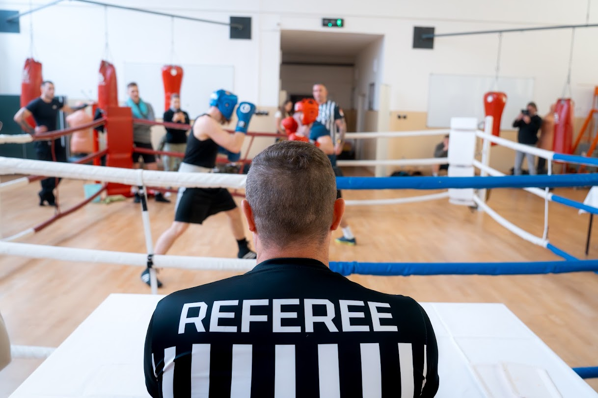 Rozhodčí sleduje zápas dvou boxerů v ochranných přilbách v ringu.